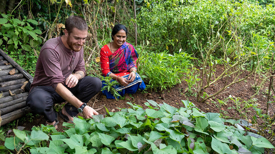 A Peace Corps volunteer kneels in front of a crop field with a woman in a sari